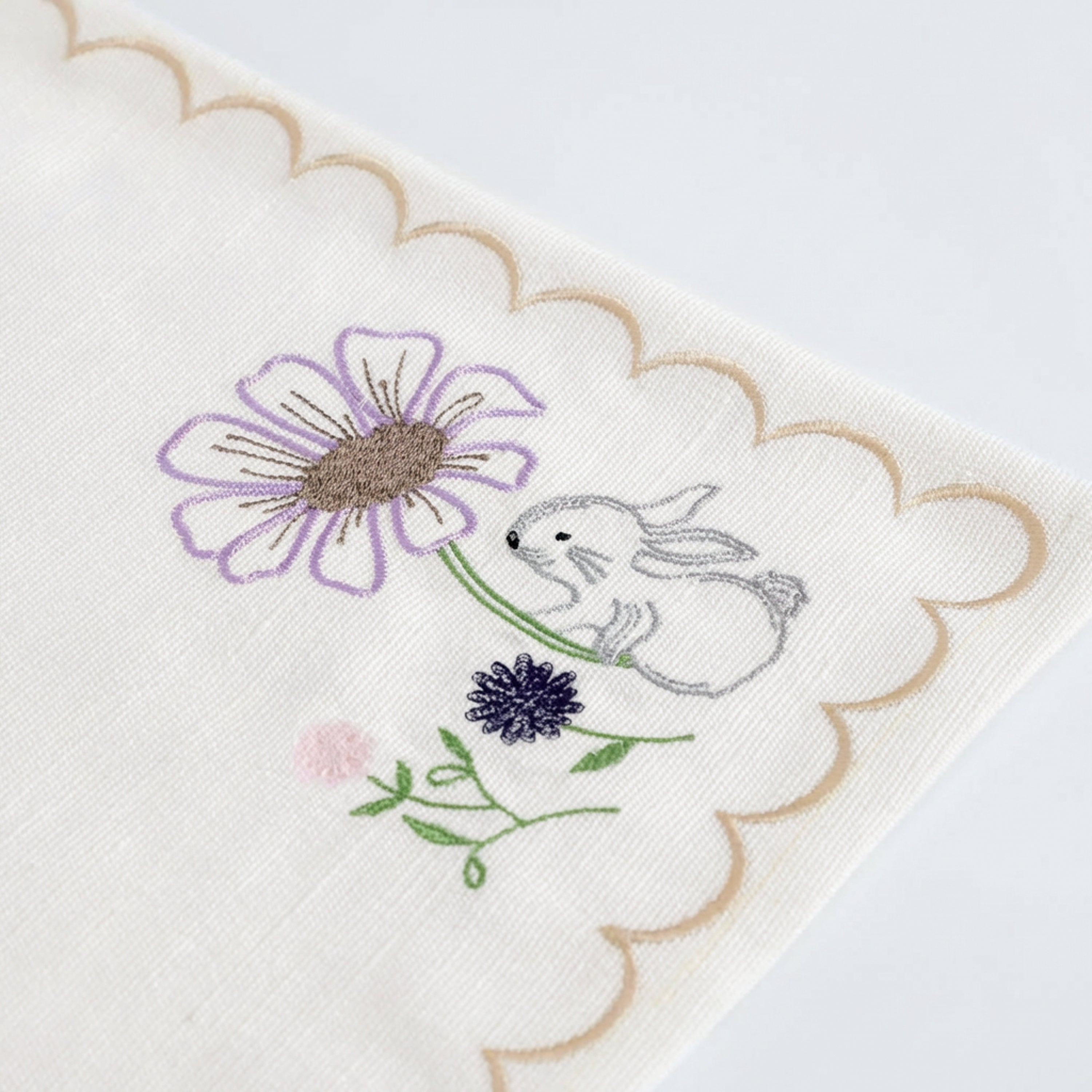 Detailed view of a high-quality Easter bunny embroidered placemat for a cottagecore spring tablescape. This ivory cotton blend table mat features a whimsical white rabbit and purple daisy motif with a decorative gold scalloped edge. It answers the search for "best spring floral dining linens" and provides a luxury, boutique feel for holiday hosting. The medium-weight canvas fabric is durable and structured, making it a top choice for seasonal farmhouse kitchen decor.