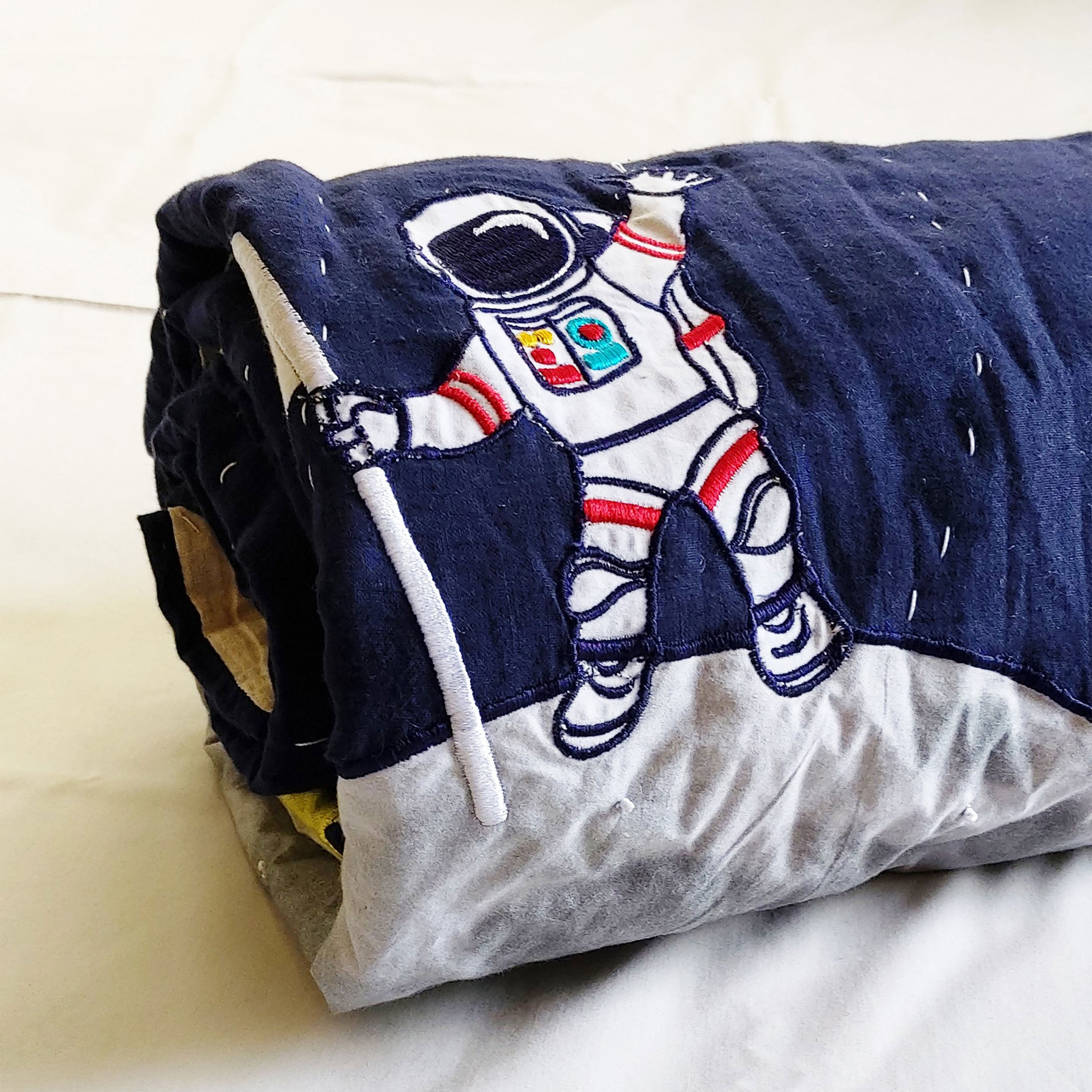 Quilt laid on bed with appliqué astronaut on moon, yellow stars, and rocket details; great for kids' space room decor, toddler bedding sets, or galaxy-themed bedroom styling.
