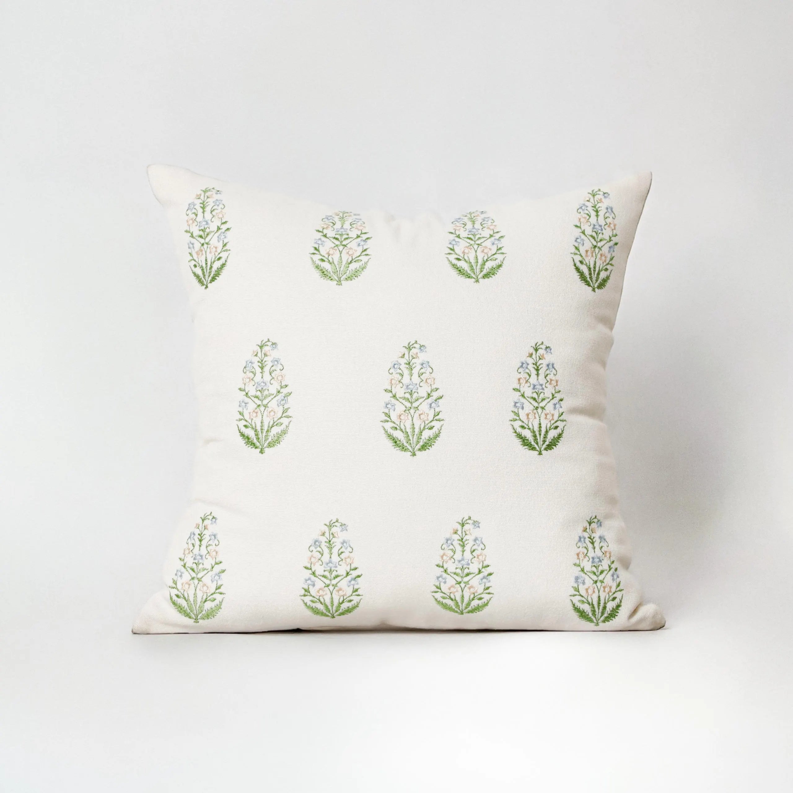 A full view of a square decorative throw pillow with a symmetrical botanical block embroider pattern, featuring green leaves and blue flowers on an off-white cotton canvas. This made-to-measure, embroidered pillow cover adds an artisanal touch to living room or bedroom decor. Ideally styled for minimalist, rustic, and European-inspired interiors, it offers a perfect balance of style and durability. A versatile accent piece for curated Airbnb, boutique hotel interiors, or a cozy, refined home environment.