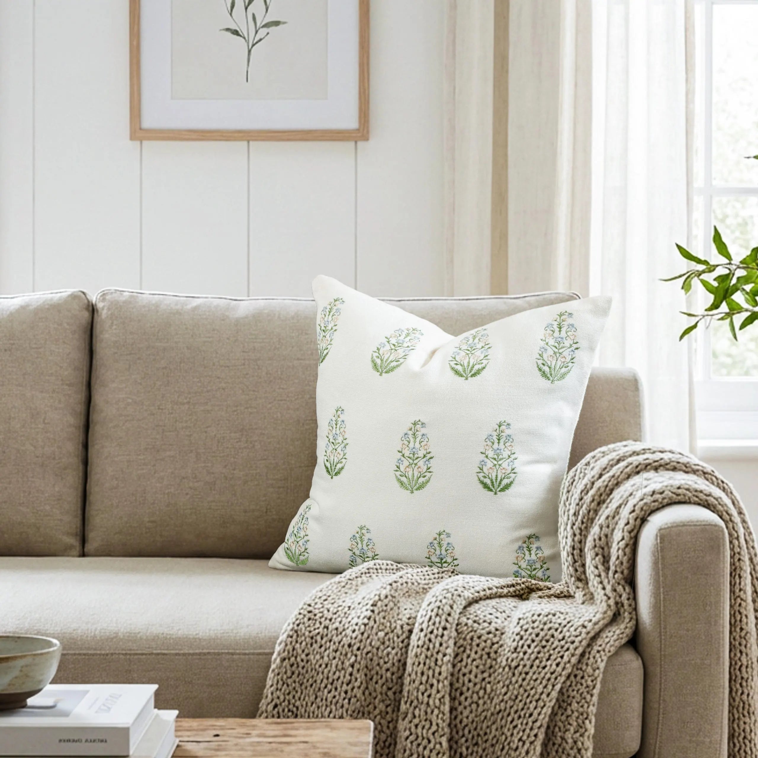 A cozy living room styling showing a neutral beige sofa with a textured knit throw blanket and a botanical block embroidered decorative pillow. The pillow features a traditional green motif on cotton canvas, illuminated by soft natural light from a window. Perfect for creating a calm, earthy, and minimal aesthetic, this setup inspires farmhouse, organic modern, or European-style interiors. Our custom-printed pillows complement made-to-measure drapery for a complete, designer home refresh.