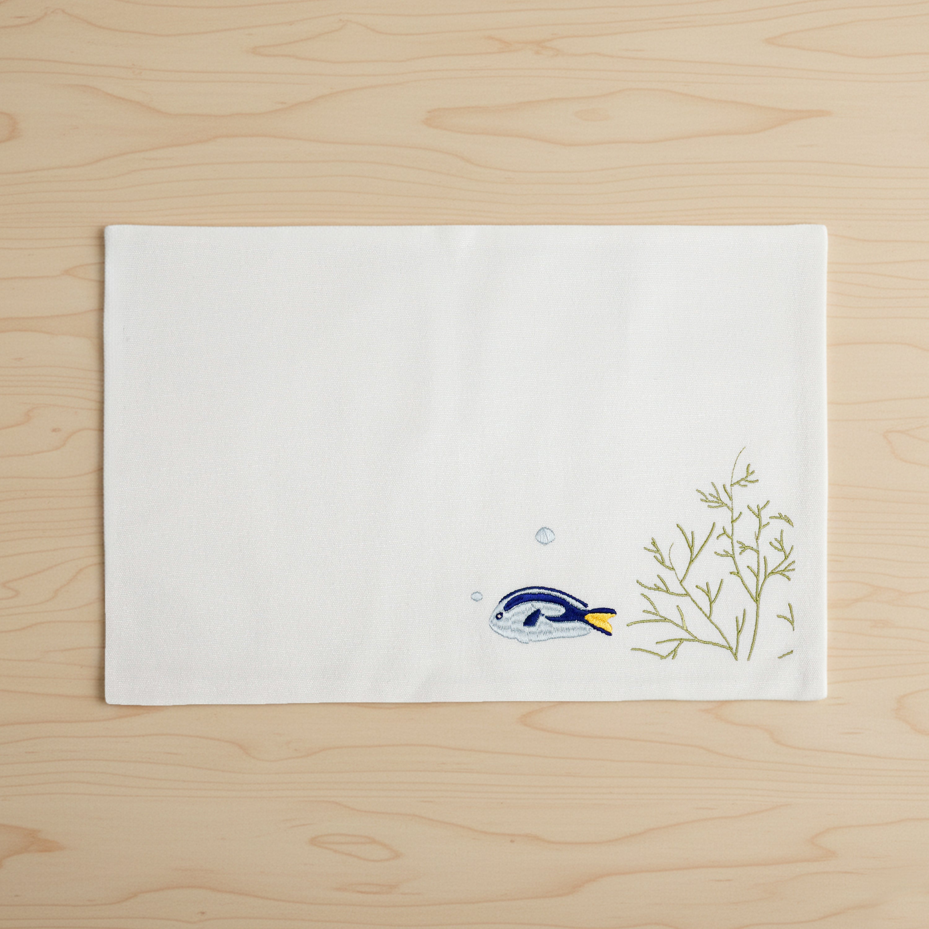 Premium ivory nautical placemats with blue tang fish embroidery for coastal dining room décor. This 12x18 inch cotton blend table mat features a modern coastal design with seaweed accents, answering the search for unique nautical-themed home accessories. Designed for durability and low maintenance, it is the top choice for families looking for high-quality, pre-washed table linens that offer a beach-inspired look for farmhouse or contemporary dining rooms.