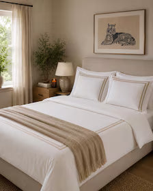 A neatly made bed in a softly lit bedroom, dressed in white bedding with a beige throw blanket draped across the foot. The room includes a nightstand, lamp, window with curtains and framed wall art above the bed.