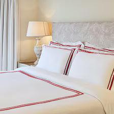 A neatly made bed styled with the 400 Thread Count Satin Cotton Duvet Cover Set with red Trim. The crisp white bedding features elegant, parallel red embroidered lines along the edges of the duvet and Four matching pillows.