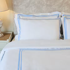 400 Thread Count Satin Cotton Duvet Cover Set with light blue Trim. The clean white bedding sets a calm tone with soft, parallel light blue embroidered borders framing the edges of the duvet and stacked pillows.