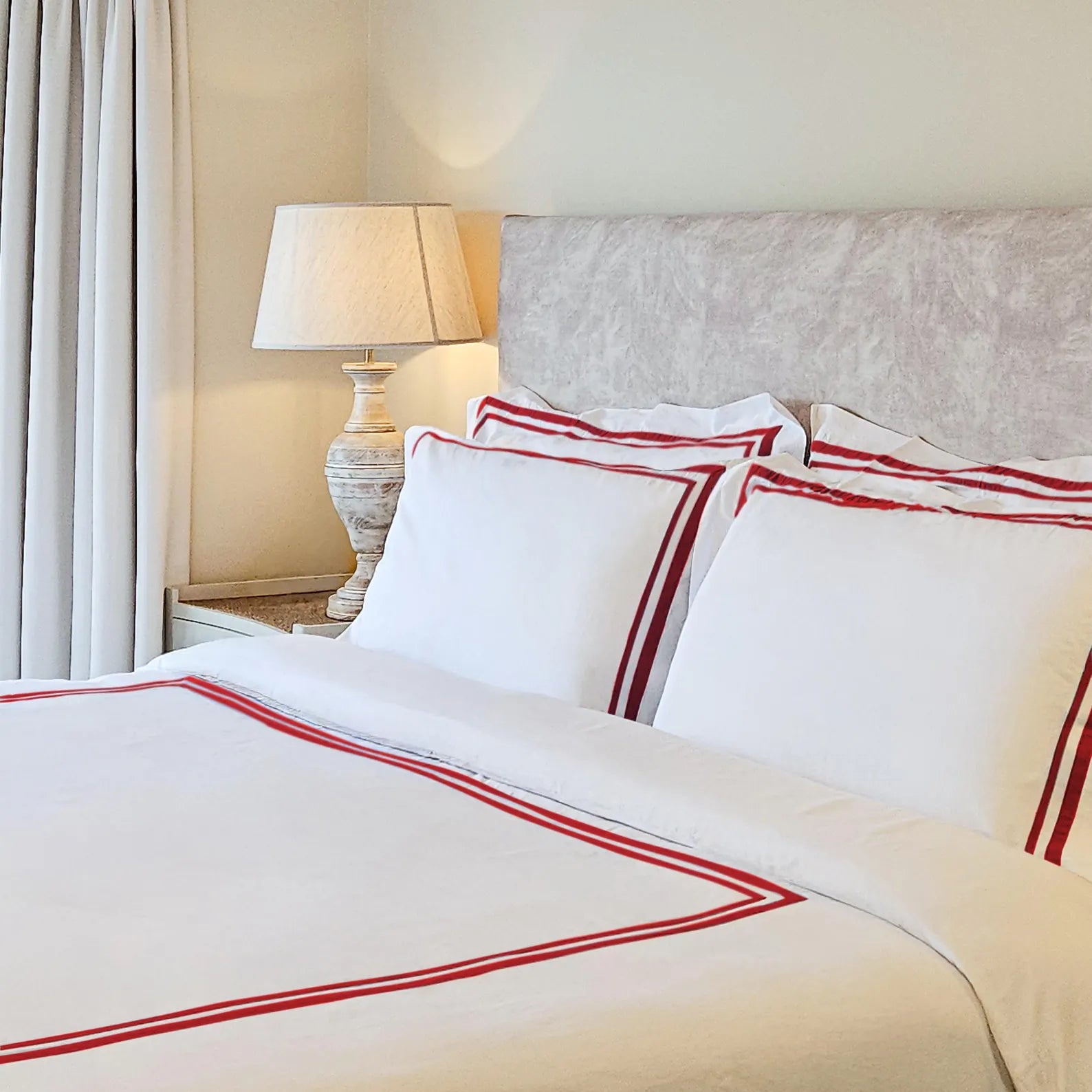 Luxury hotel bedding by Amore Beauté: custom trimmed duvet cover set and matching pillow shams in crisp white 400TC cotton blend. Features unique double red tailored stripes, meticulously detailed in this styled bedroom shot with a textured headboard and a decorative lamp on a nightstand. This sophisticated, personalized set can be custom-made in bespoke sizes to perfectly fit homes, premium hotels, and luxury spas seeking high-end decor.