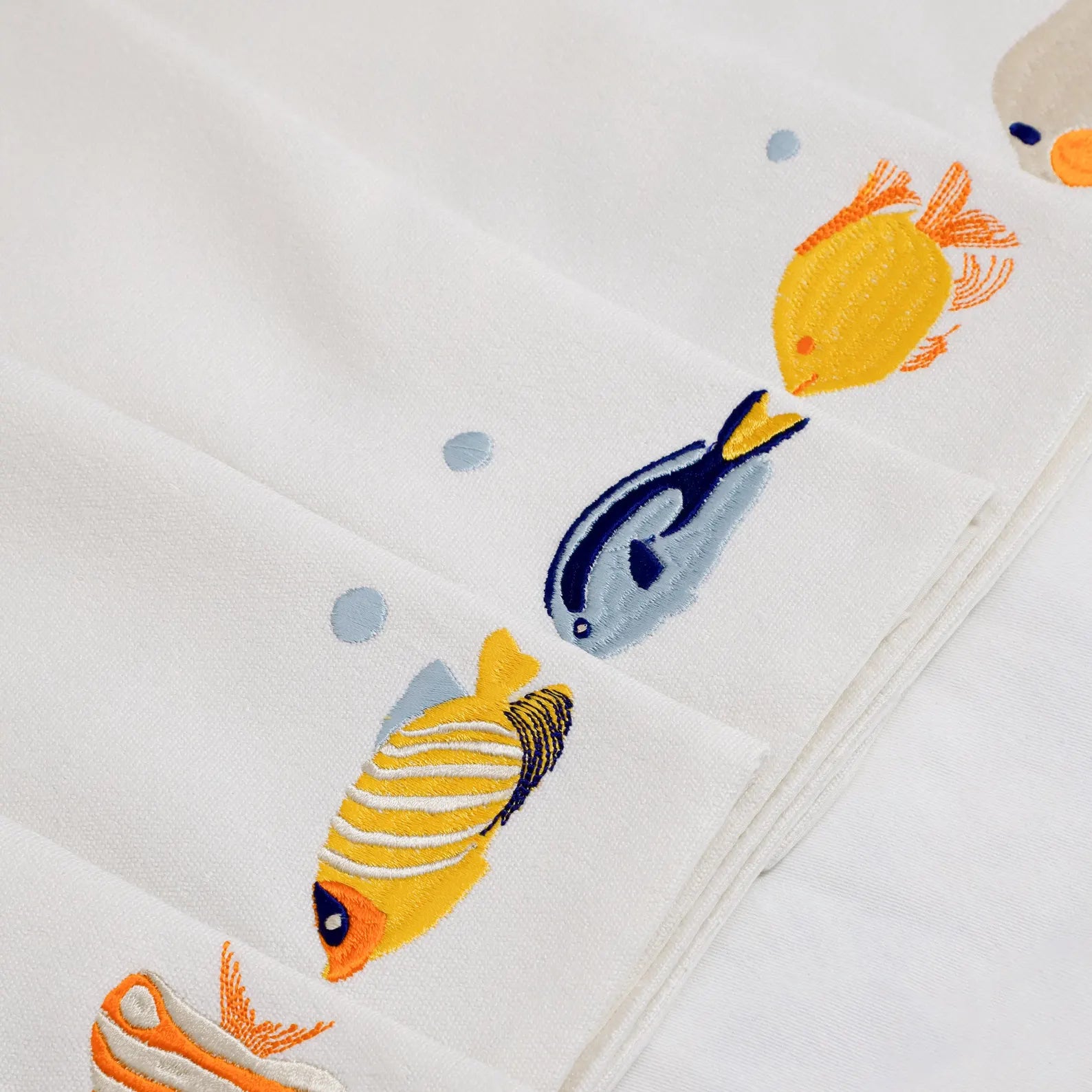 How to style a nautical-themed table for summer entertaining with embroidered fish placemats. This close-up view displays a set of 6 premium ivory cotton placemats, each featuring different vibrant sea life embroidery, including yellow and blue tropical fish. The soft cotton backing provides extra structure, making these the best luxury coastal table linens for formal occasions or casual seaside meals. These handmade, easy-care placemats are an essential gift for beach house owners.