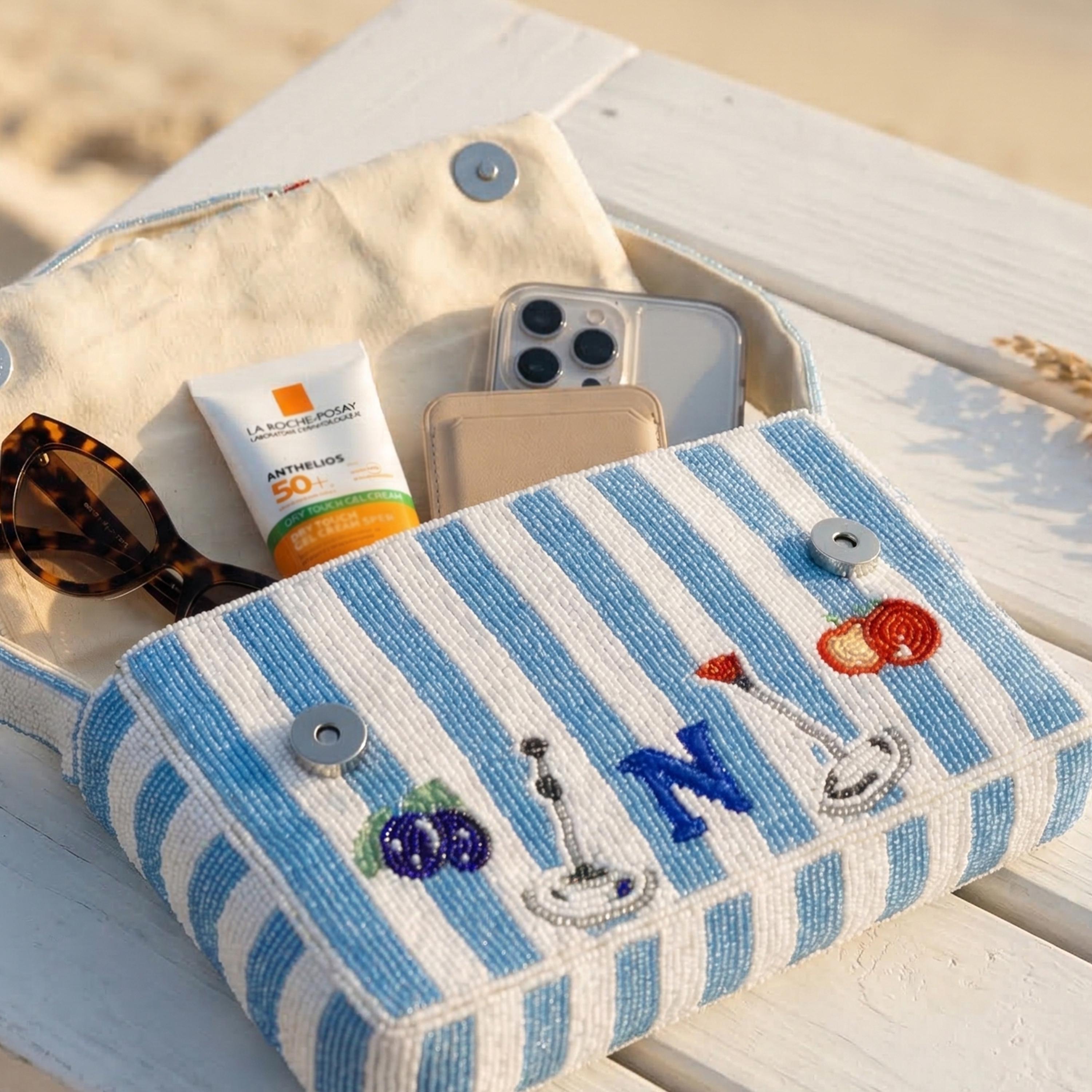 Open hand-beaded mini shoulder bag in blue and white stripes by Amore Beauté, showing the contents: sunglasses, phone, cardholder, and SPF. This luxury handmade purse features playful cocktail and fruit beadwork with a personalized monogram 'N'. An essential summer vacation accessory for women, it’s designed to hold brunch and beach party necessities. The magnetic snap closure ensures security. A perfect, unique statement piece for a playful luxury aesthetic and personalized gifting.